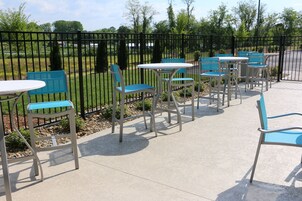 An outdoor patio area with tables and chairs, a metal fence, and a view of a park with trees and a road.