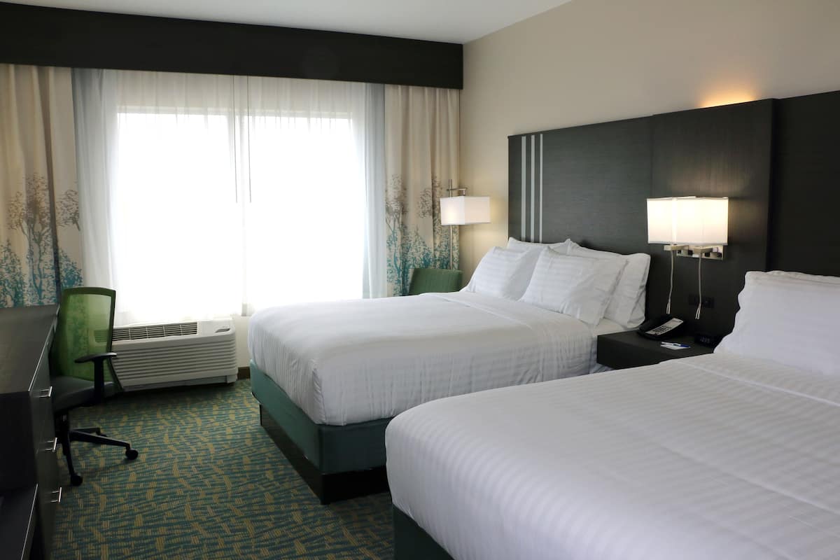 A hotel room with two beds, a desk, a chair, a lamp, and a window with curtains.