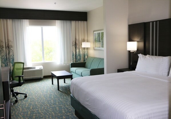 Quarto de hotel com uma cama grande, um sofá verde, uma mesinha, uma escrivaninha e uma janela com cortinas.