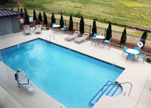 A rectangular swimming pool with a tiled edge, surrounded by lounge chairs and tables, and a fenced area with trees. - Holiday Inn Express & Suites Hendersonville SE - Flat Rock by IHG (Flat Rock)