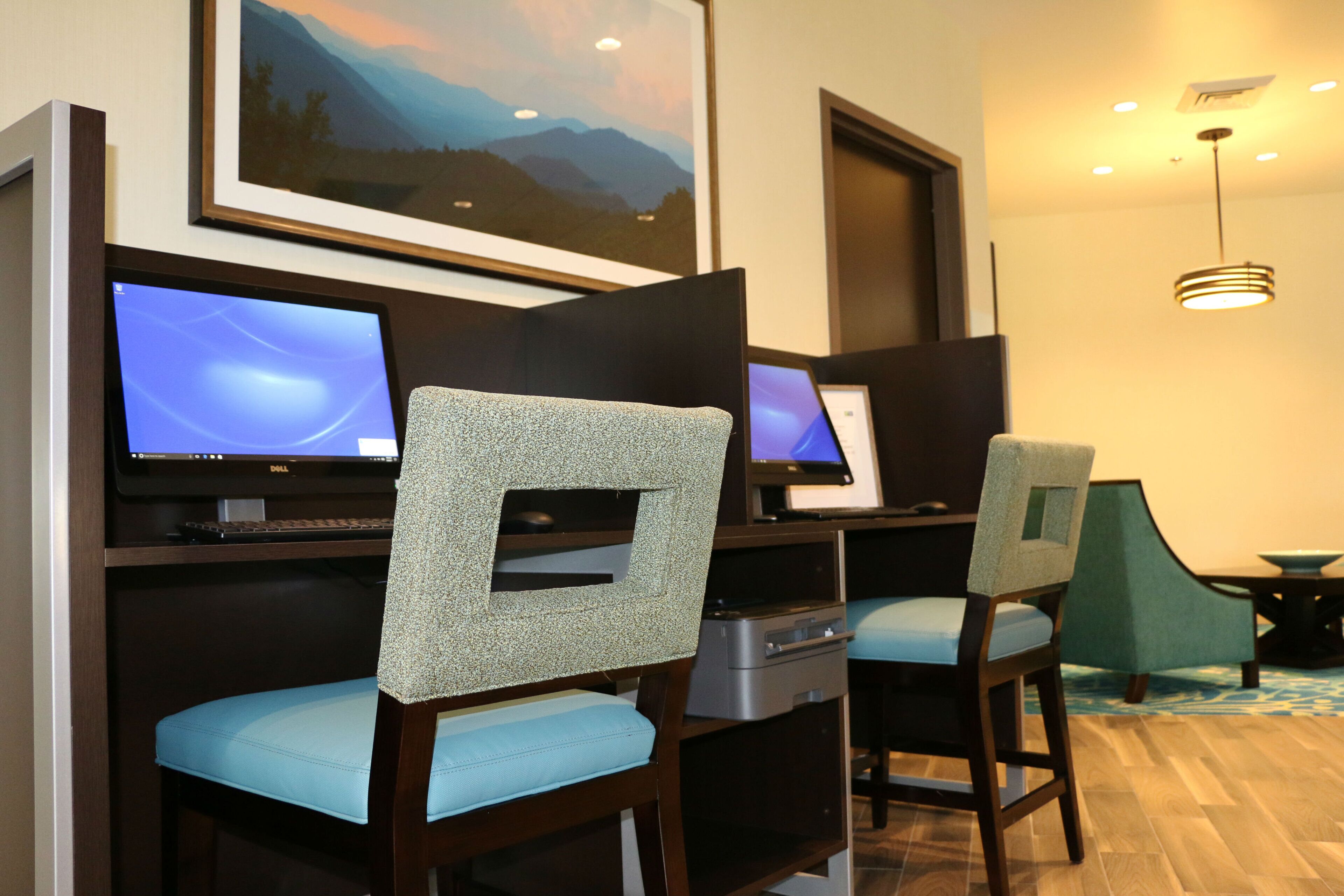 Une chambre d’hôtel avec deux bureaux, chacun équipé d ’un écran d’ordinateur, d’un clavier et d’une souris. Il y a deux chaises avec des coussins bleu clair. Un tableau représentant des montagnes est accroché au mur, au-dessus des bureaux.