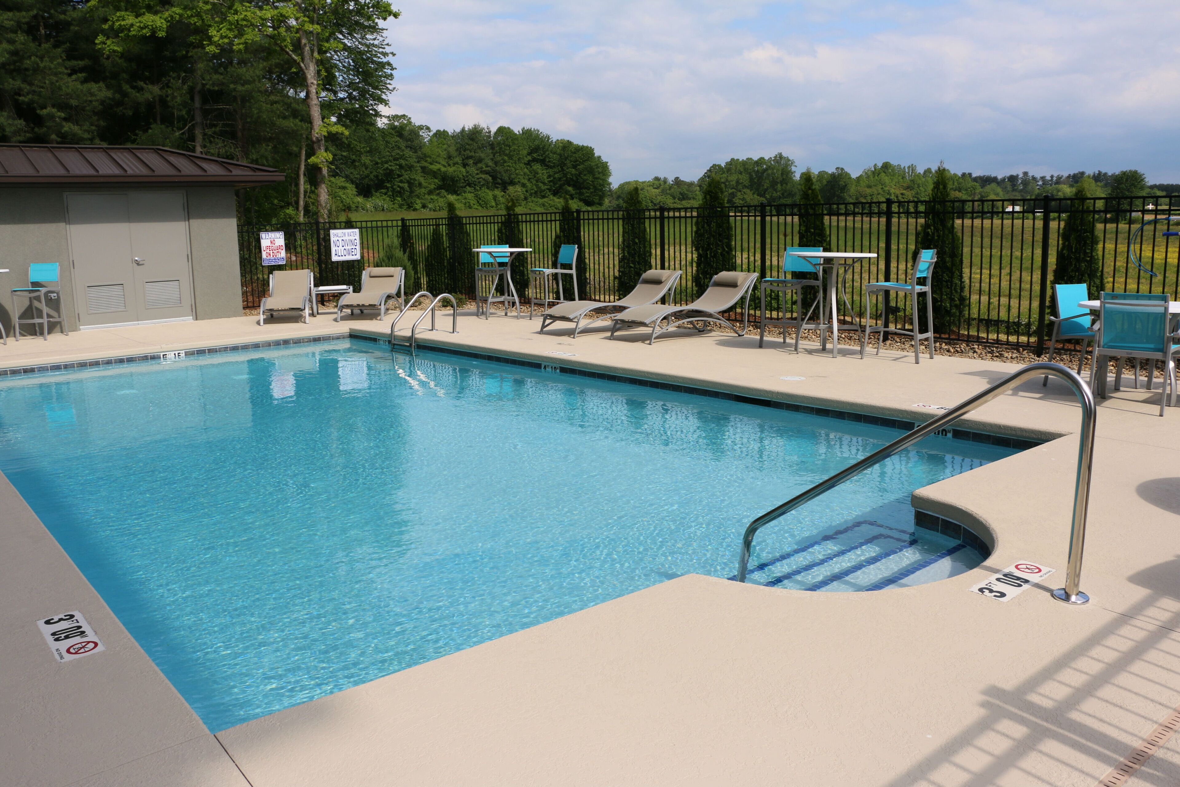 A swimming pool with lounge chairs and tables, surrounded by a fence and trees.