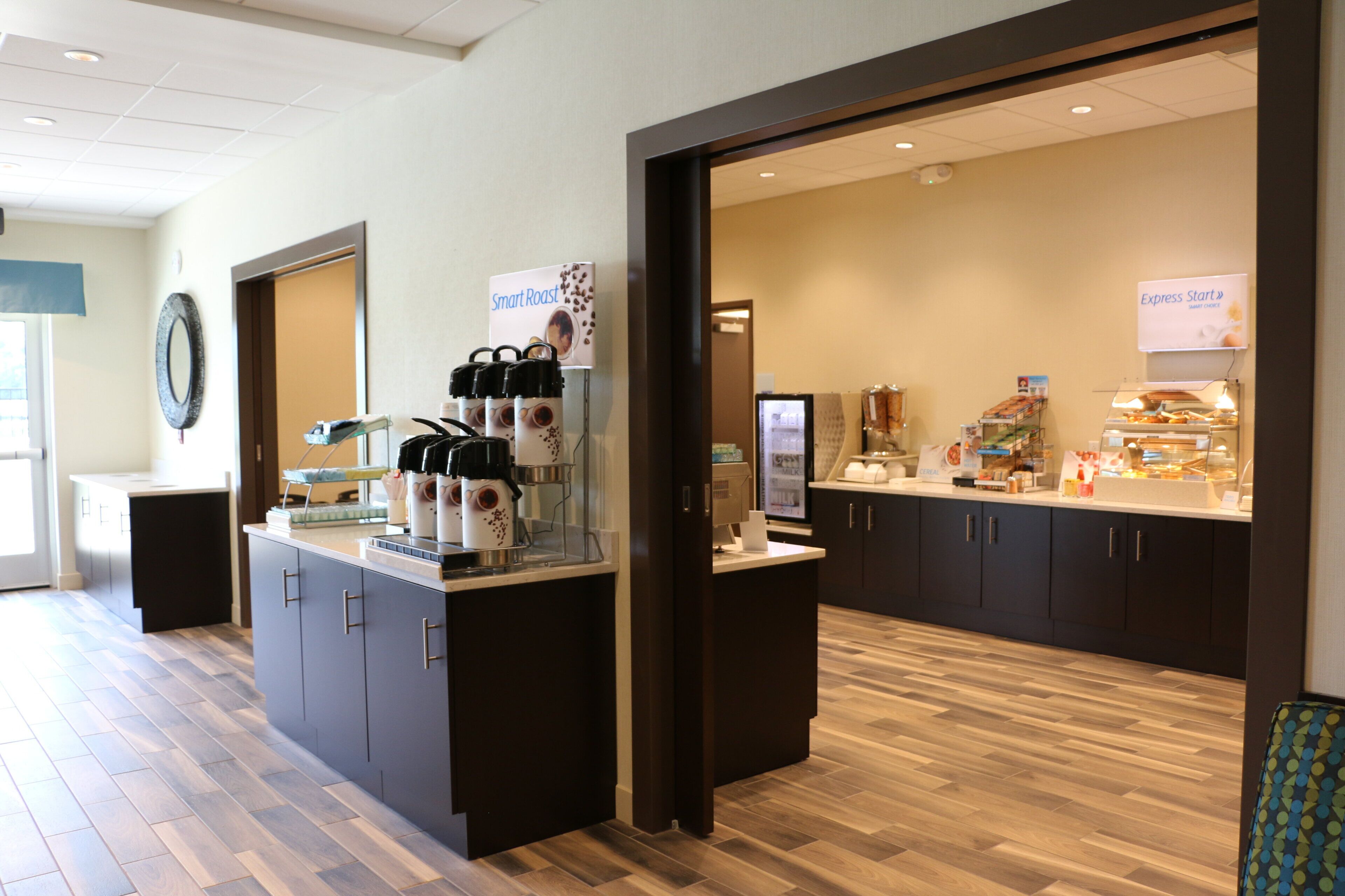 A hotel breakfast area with a coffee station, a refrigerator, and a display of various food items.