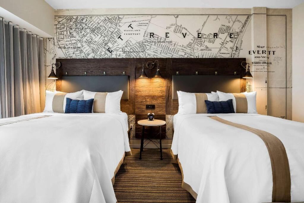 Quarto de hotel com duas camas, uma cabeceira de madeira, um mapa na parede e uma pequena mesa com um relógio.