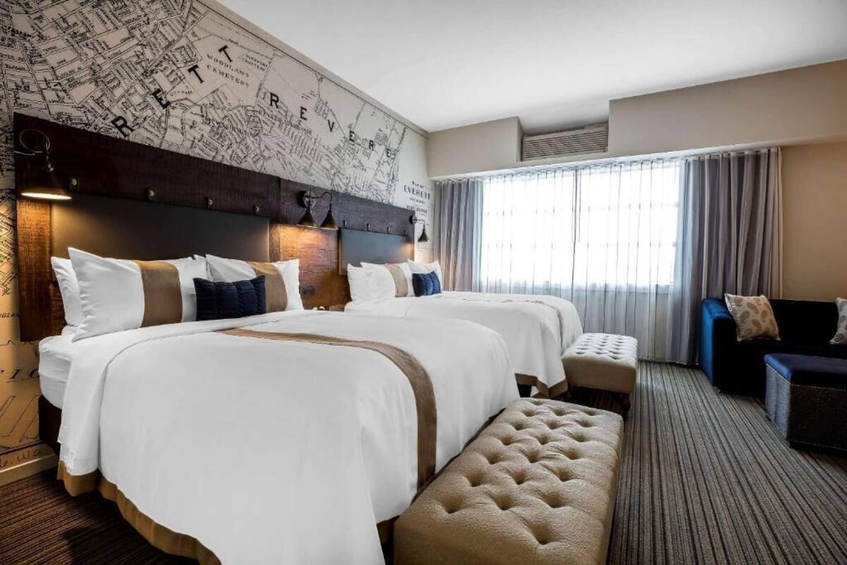 Une chambre d’hôtel avec trois lits, une grande carte accrochée au mur, une fenêtre avec des rideaux et un canapé de couleur sombre.