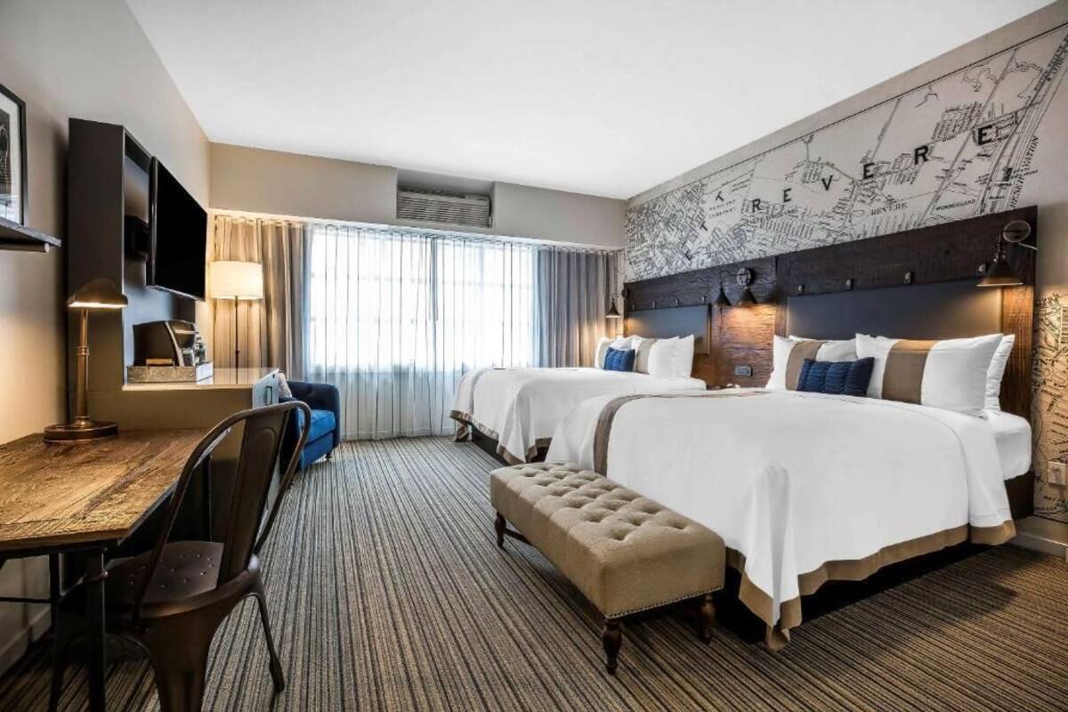 Une chambre d’hôtel avec deux lits, un bureau, une chaise, une télévision et une grande fenêtre avec des rideaux.