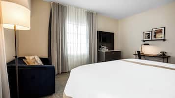 A hotel room with a large bed, a desk, a chair, a sofa, a TV, and a lamp.
