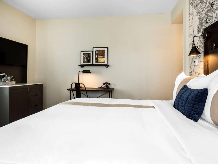 A hotel room with a bed, a desk, a chair, a lamp, and two framed pictures on the wall.