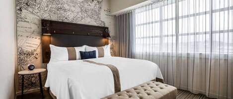 Une chambre d’hôtel avec un grand lit, une tête de lit en bois, une carte accrochée au mur, une petite table et une grande fenêtre avec des rideaux.