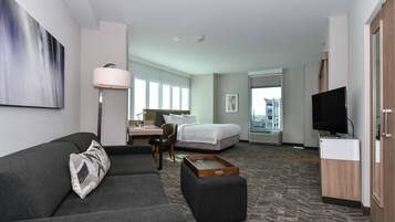 Une chambre d’hôtel moderne avec un canapé gris, une table d’appoint en bois, un lit, un bureau avec une chaise, une télévision et une grande fenêtre donnant sur la ville.