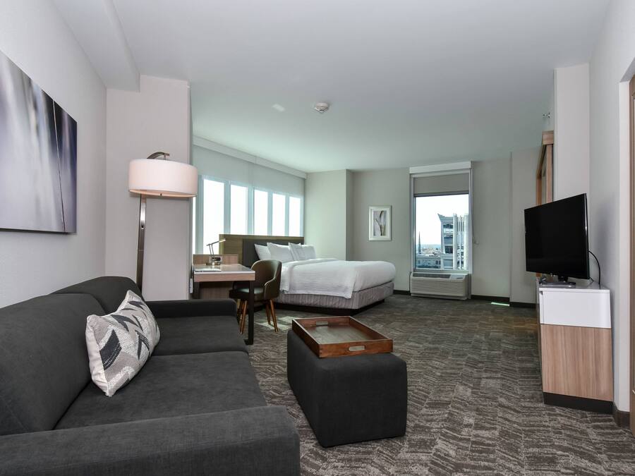 A modern hotel room with a gray sofa, a wooden side table, a bed, a desk with a chair, a TV, and a large window offering a city view.