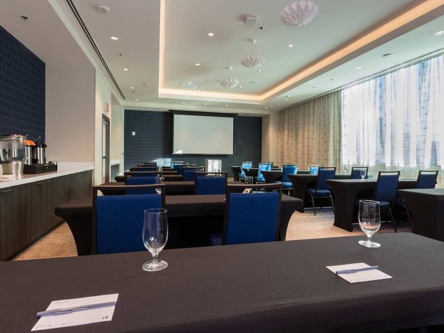 Un salón de conferencias con mesas, sillas, una pantalla de proyector y una zona de cocina con electrodomésticos.