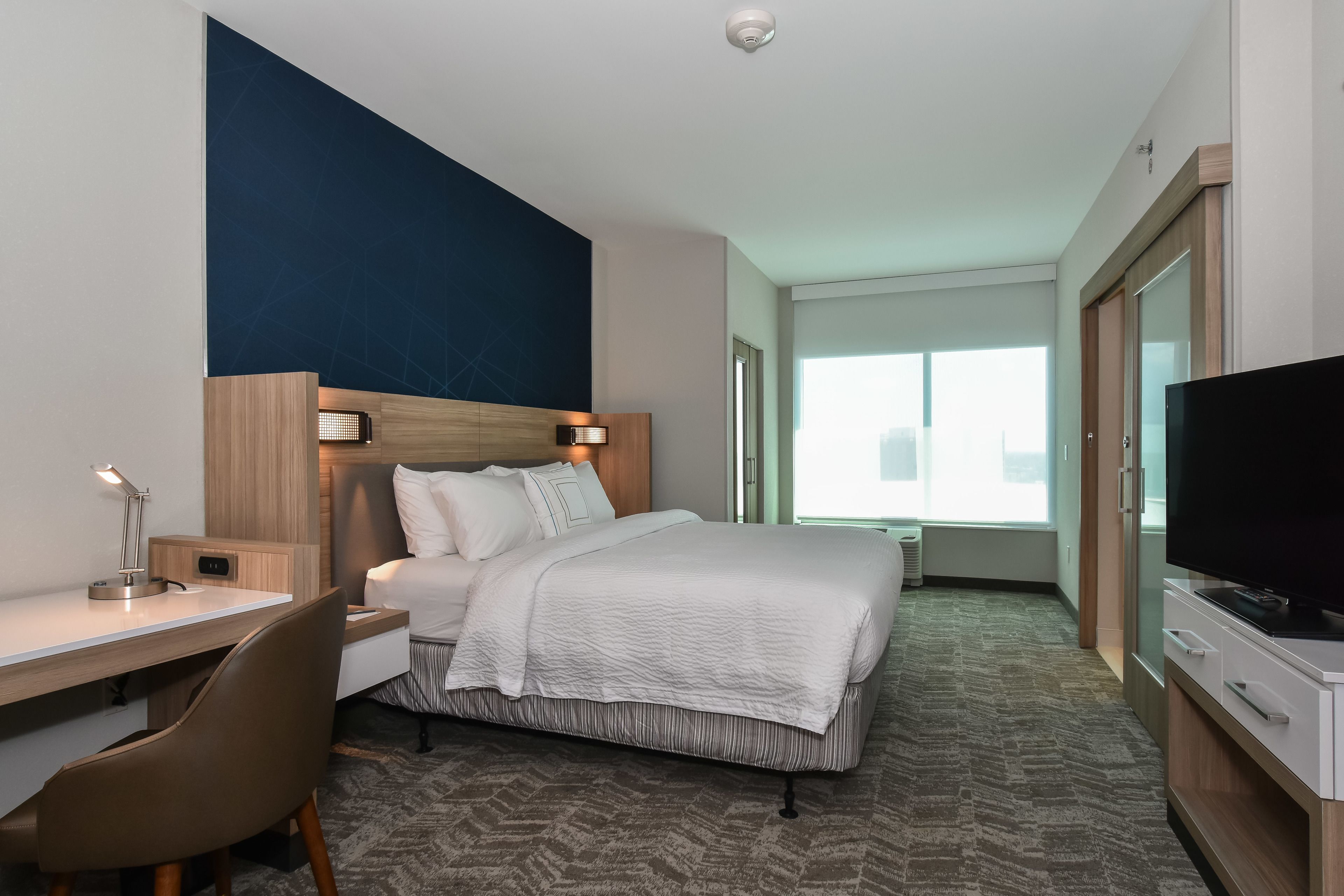 Ein Hotelzimmer mit einem Bett, einem Schreibtisch mit Stuhl, einem Fernseher und einem großen Fenster.