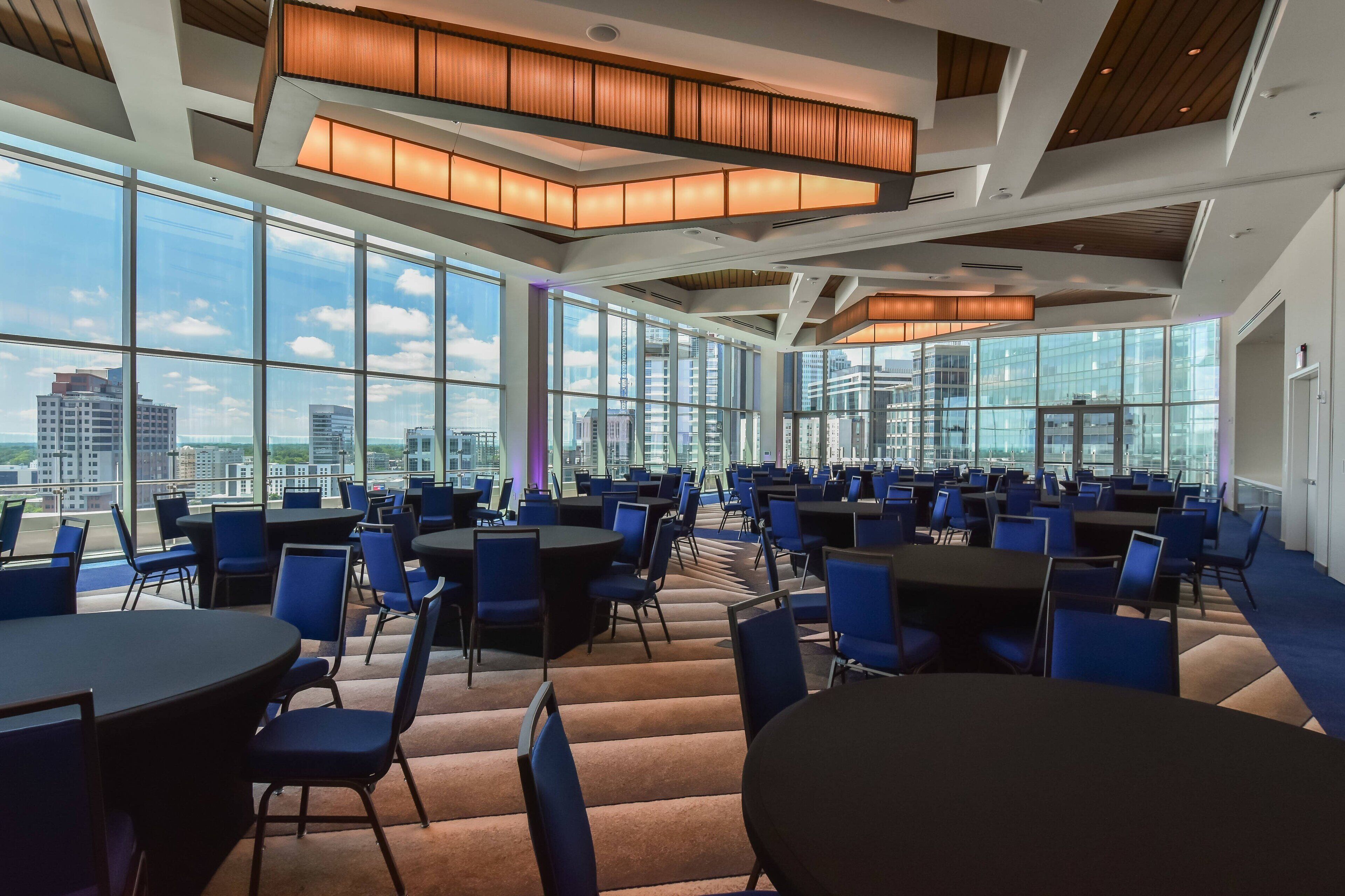 A spacious conference room with multiple tables and chairs, large windows offering city views, and modern lighting fixtures.