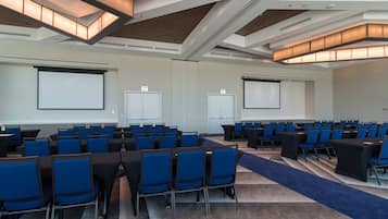 A conference room with rows of blue chairs, tables, and a carpeted floor.