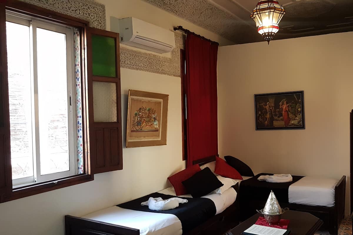 A hotel room with two beds, a large window, a wall-mounted air conditioner, a decorative lamp, and a painting.