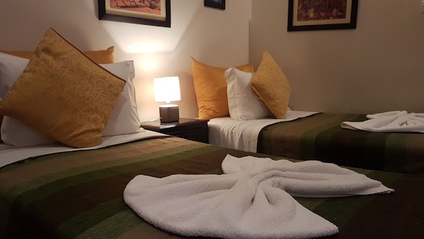 Habitación de hotel con dos camas, cada una con colcha estampada, almohadas y una manta. Hay una mesita de noche con una lámpara y un cuadro en la pared.