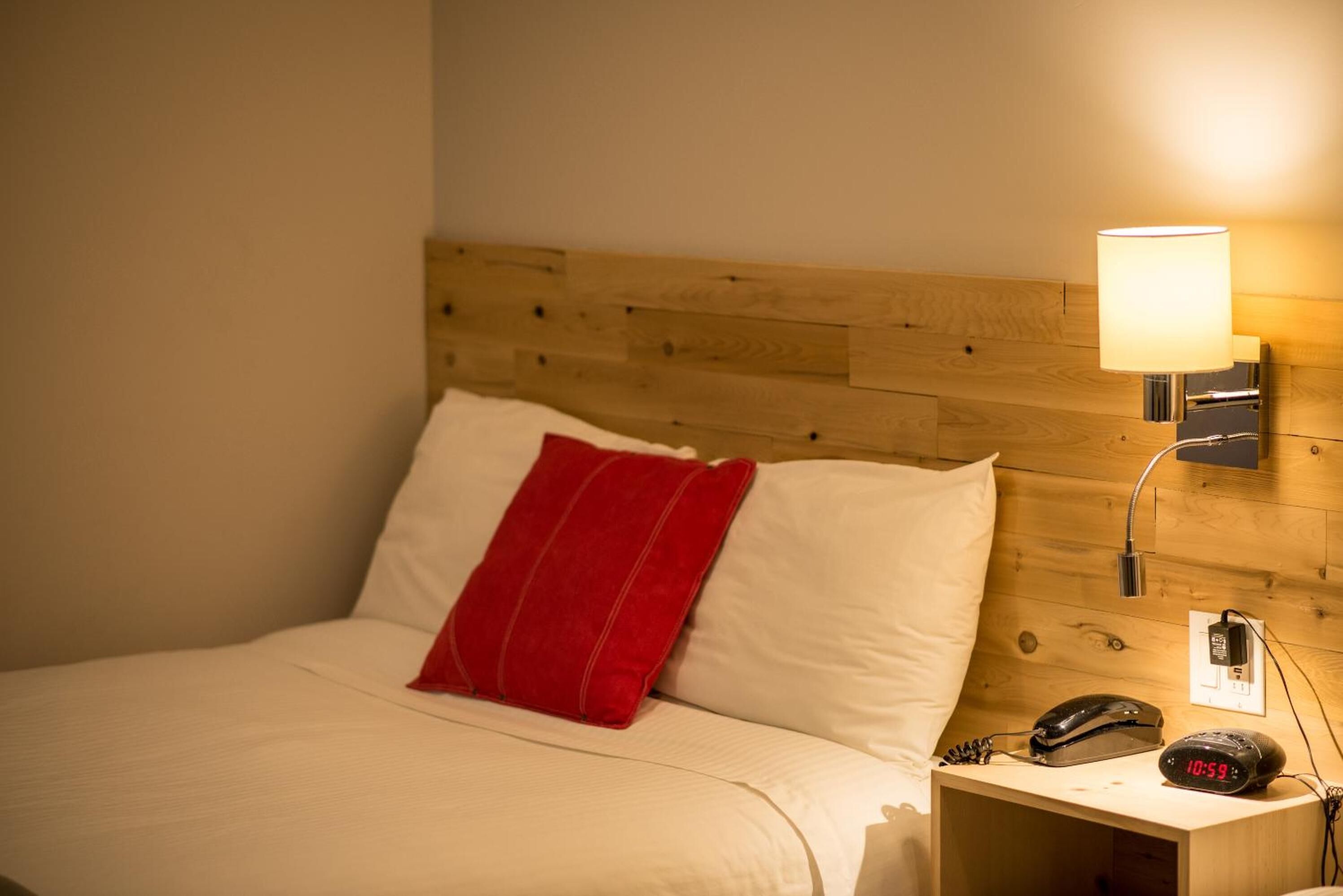 A hotel room with a wooden headboard, a bed with white bedding and a red pillow, a bedside table with a lamp, a phone, and a digital clock displaying the time.