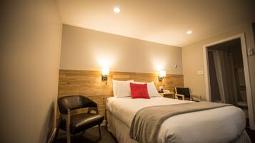 A hotel room with a wooden headboard, a bed with white bedding and a red pillow, a black leather chair, a bedside table with a lamp, and a door leading to a bathroom.