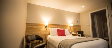 Habitación de hotel con cabecera de madera, cama con ropa blanca y almohada roja, sillón de cuero negro, mesita de noche con lámpara y puerta que da a un baño.
