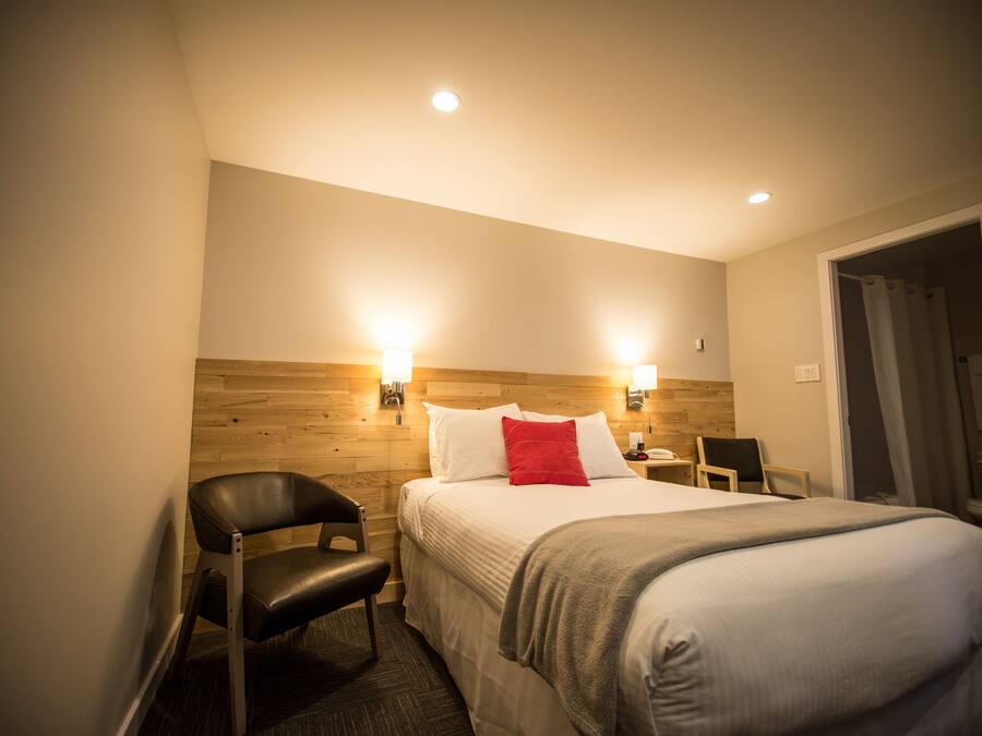 A hotel room with a wooden headboard, a bed with white bedding and a red pillow, a black leather chair, a bedside table with a lamp, and a door leading to a bathroom.