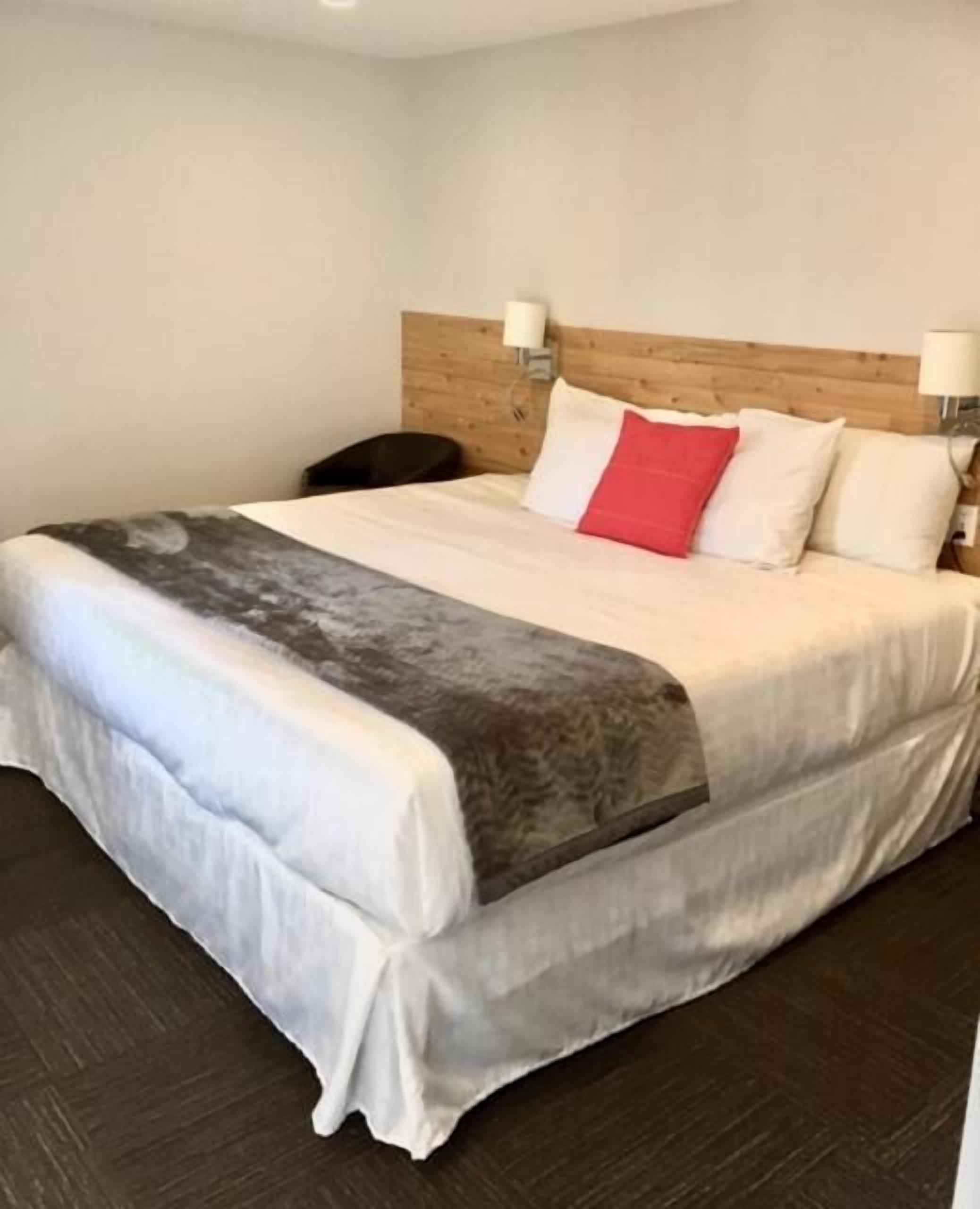 Un lit bien fait, avec une couette grise et blanche, un coussin rouge qui apporte une touche de couleur, et deux lampes fixées au mur.