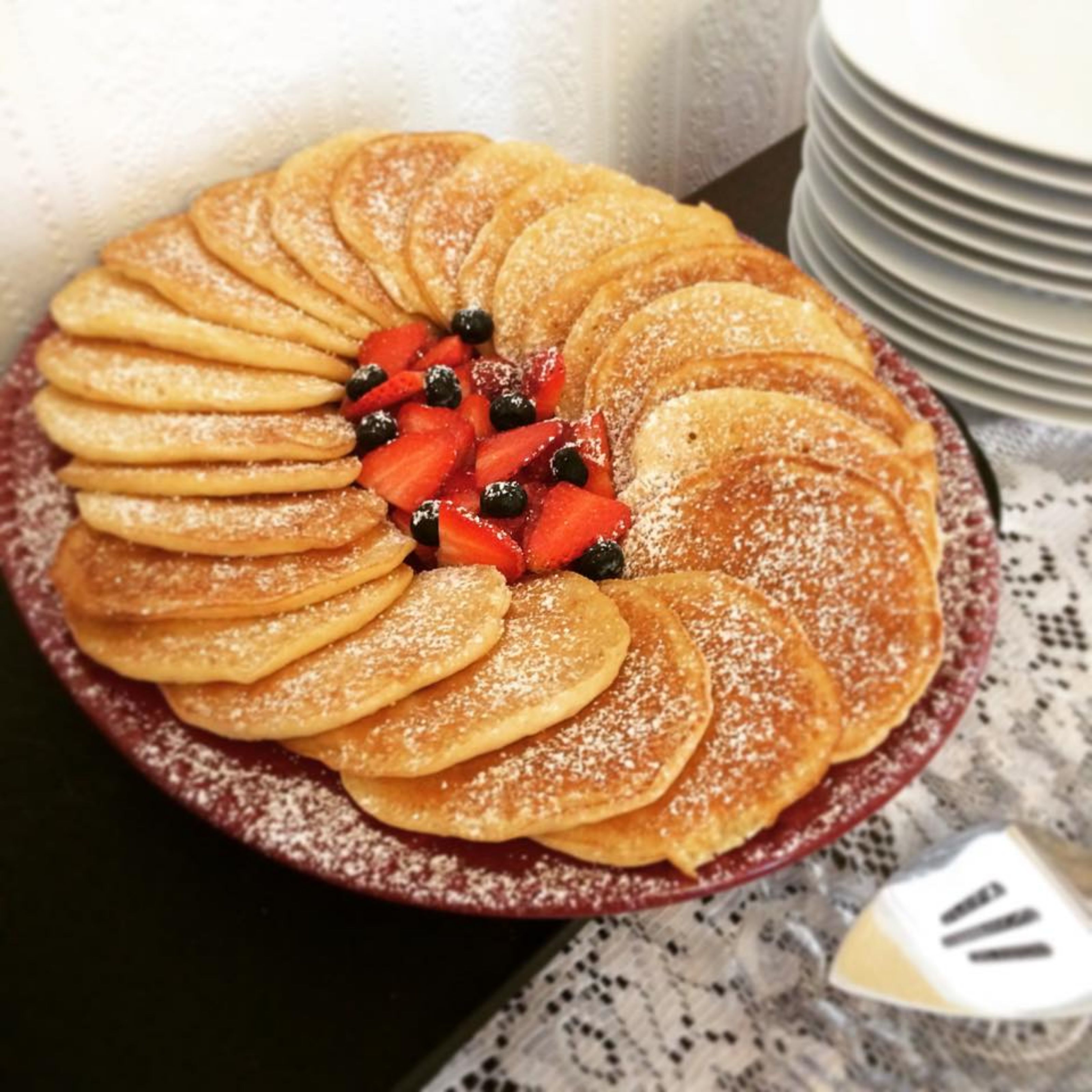 Un plato de tortitas dispuestas en forma de círculo, cubiertas con fresas y arándanos, y espolvoreadas con azúcar glas.
