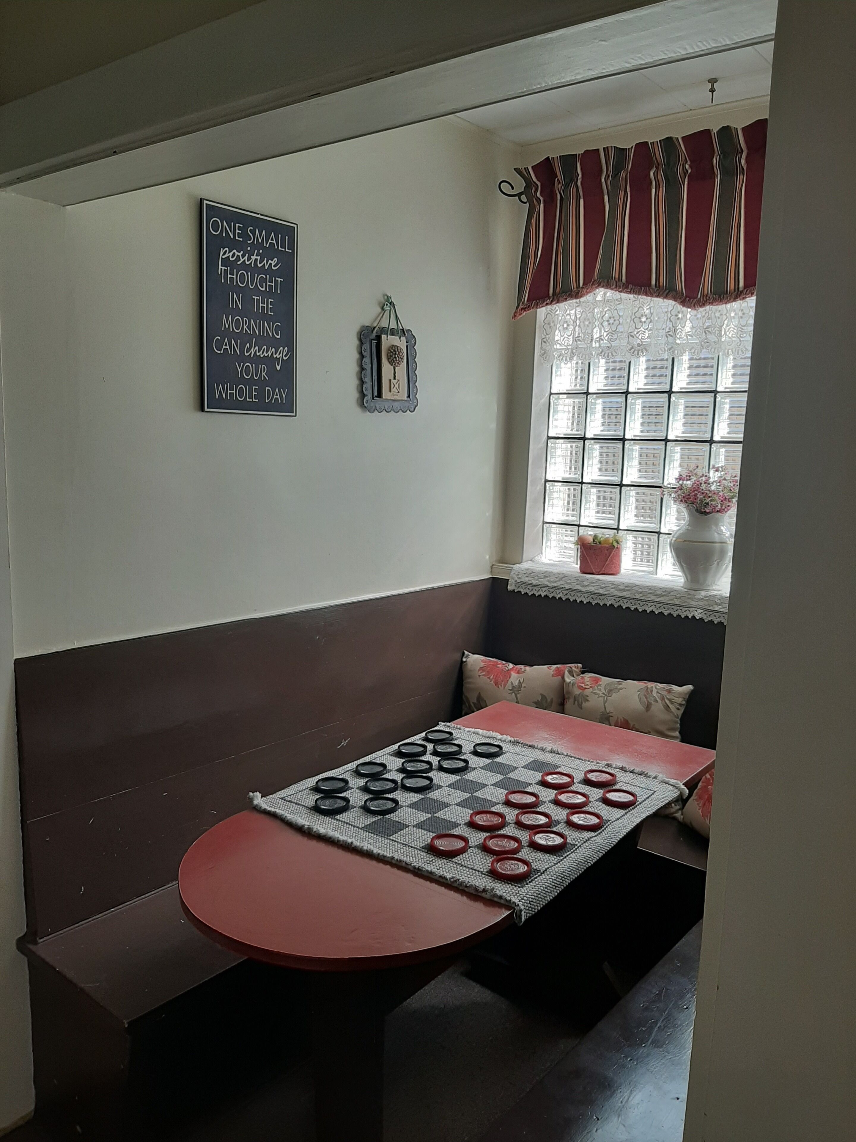 A cozy seating area with a bench and a small table, a decorative wall hanging, a window with curtains, and a potted plant.