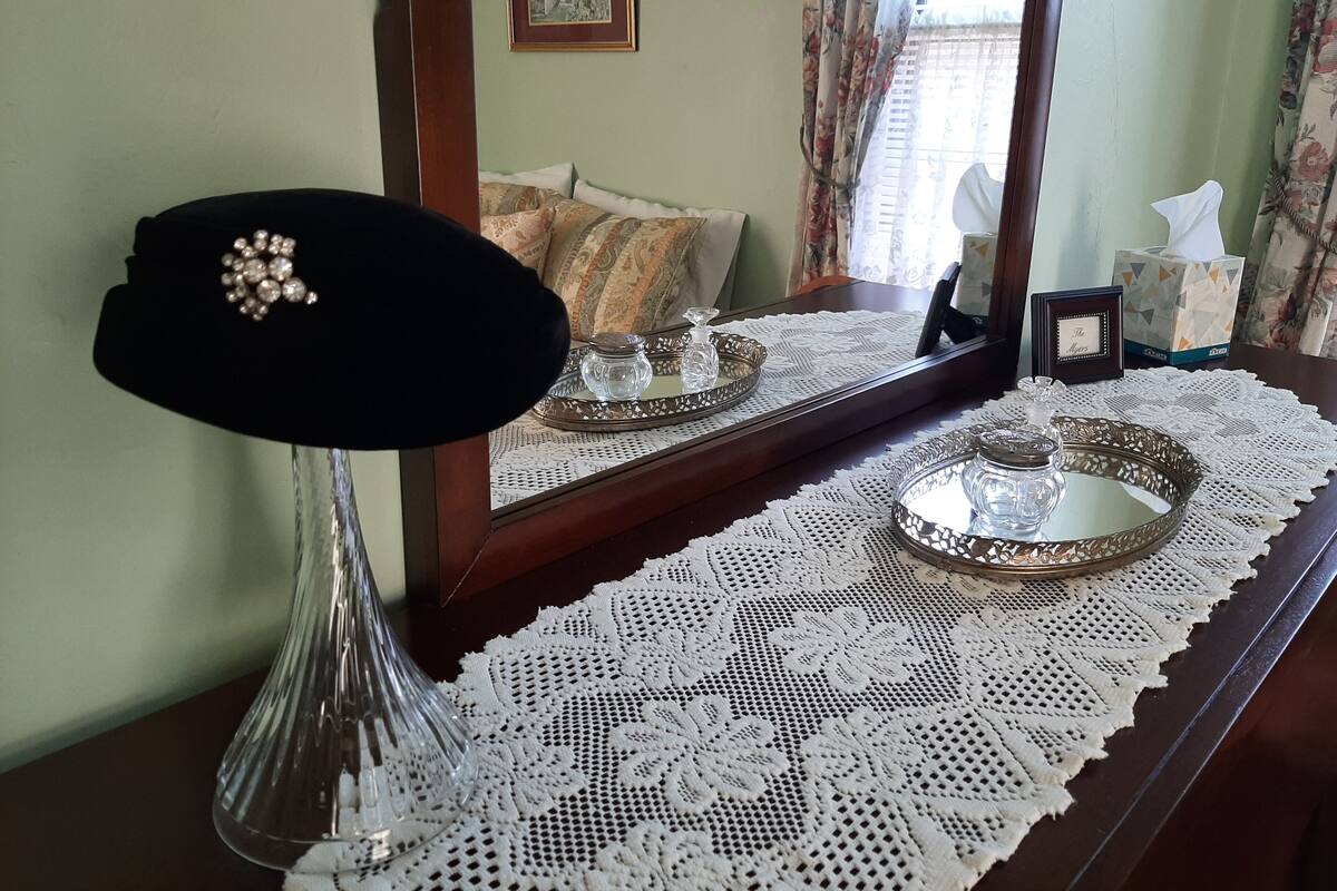 A room with a mirror, a lace tablecloth, a black hat, and a tray with a glass jar.