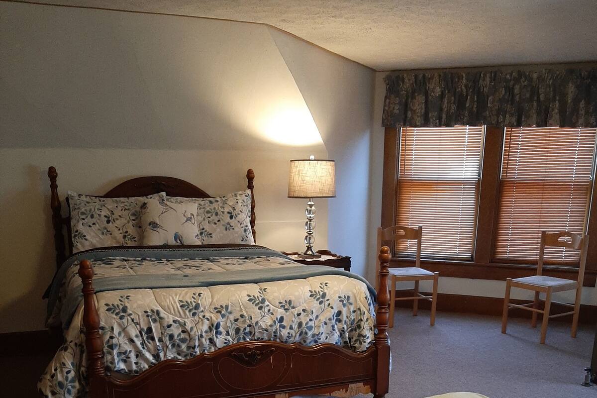 A bedroom with a large bed, a bedside table with a lamp, a chair, and a window with blinds.