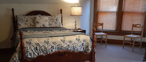 A bedroom with a large bed, a bedside table with a lamp, a chair, and a window with blinds.