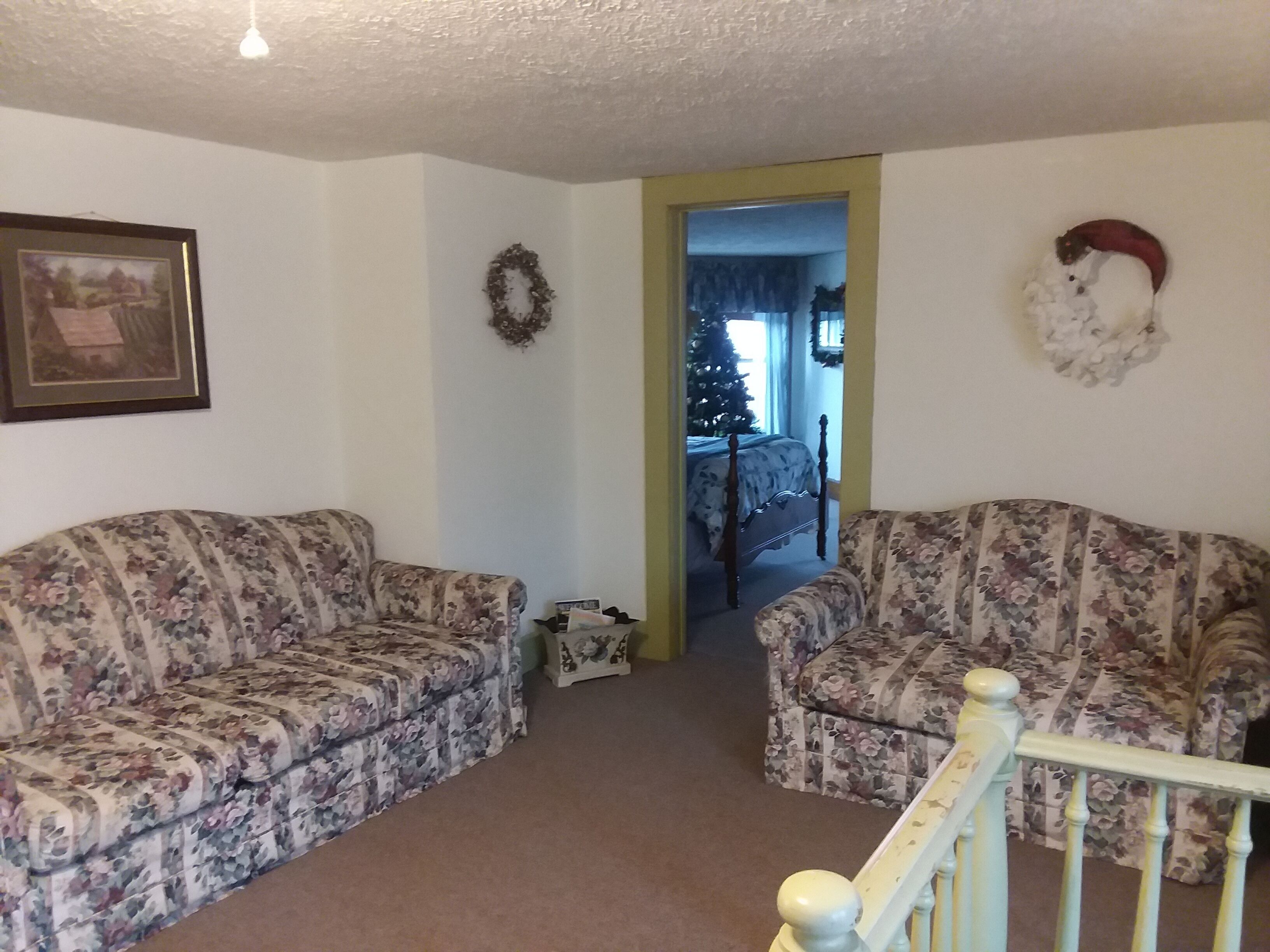 A living room with two floral-patterned sofas, a framed picture on the wall, a wreath, and a Christmas tree in the background.