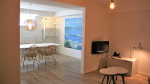 A modern dining area with a white table and wicker chairs, a painting of a seascape on the wall, and a TV on a white cabinet. - Boulevard 5 (Zandvoort)