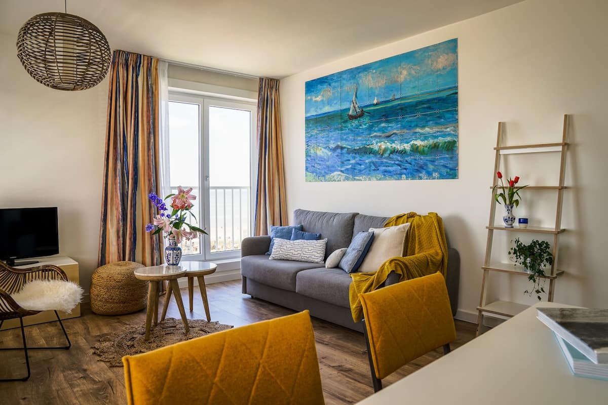 Una sala de estar moderna con un sofá gris, sillas amarillas, un televisor y un cuadro de un paisaje marino.