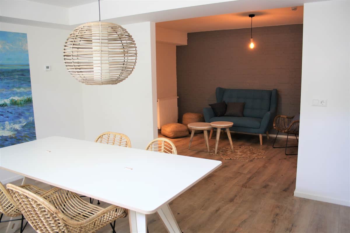 Una sala moderna con una mesa blanca, sillas de mimbre, un sofá azul y una lámpara colgante.
