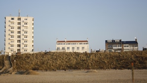 A coastal hotel with a sandy beach, a wooden staircase leading to the entrance, and a grassy dune in the foreground. - Boulevard 5 (Zandvoort)