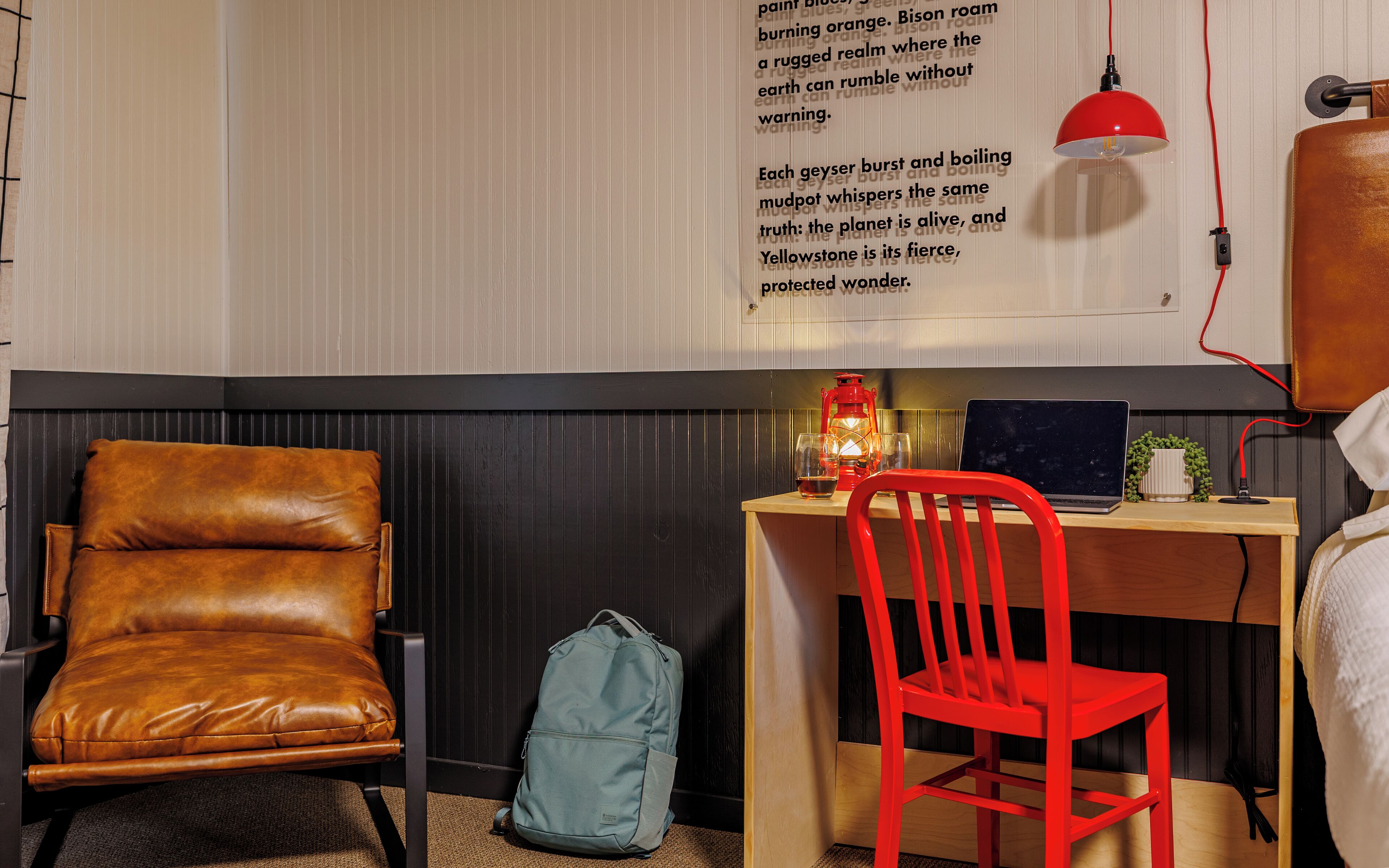 Una habitación con un escritorio de madera, una computadora portátil, una silla roja, un sillón de cuero marrón, una lámpara colgante roja y una pared con texto.