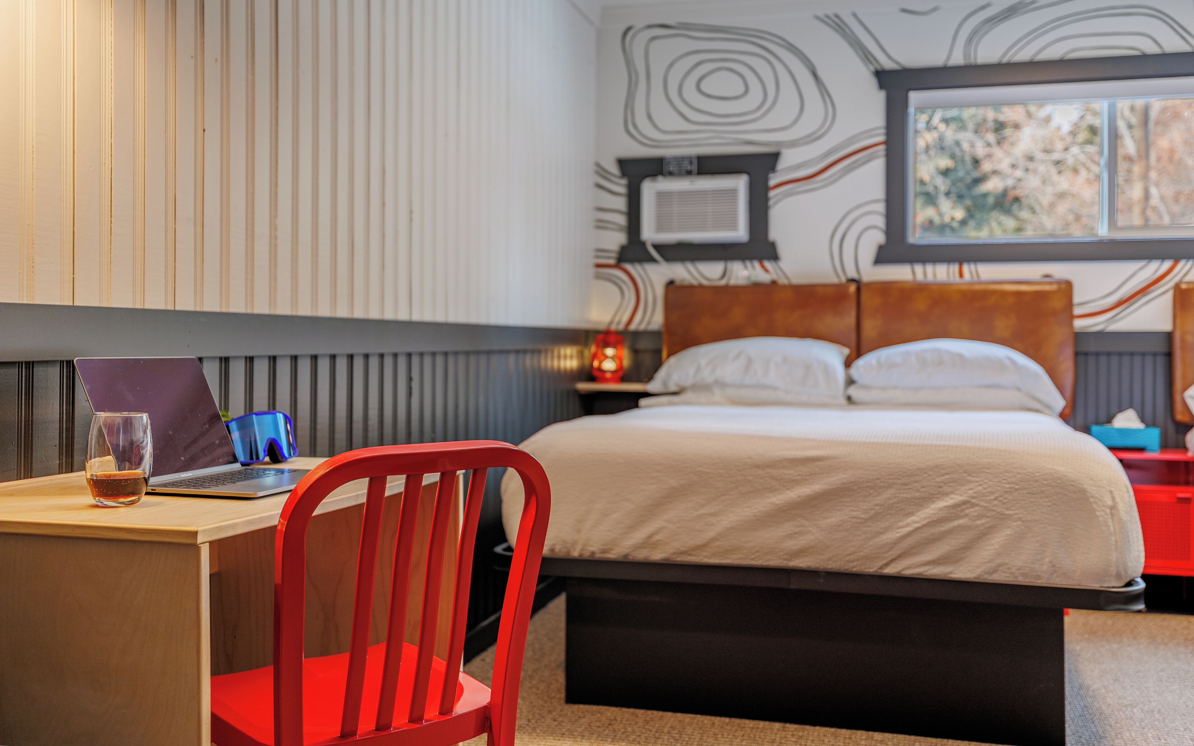 A hotel room with a bed, a desk with a laptop, a chair, and a bedside table with a lamp.