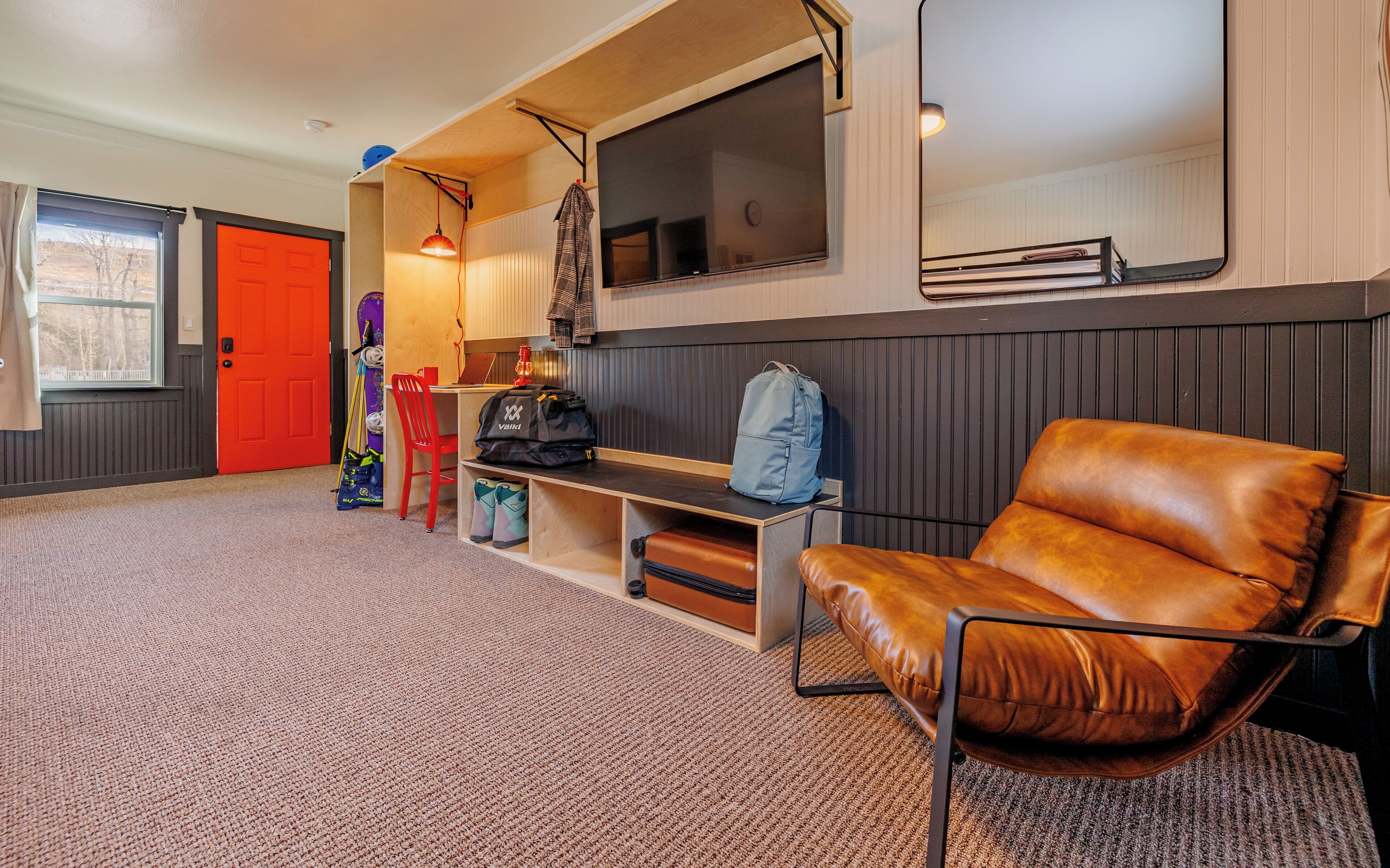 A cozy room with a leather chair, a desk with a laptop, a large window, and a red door.