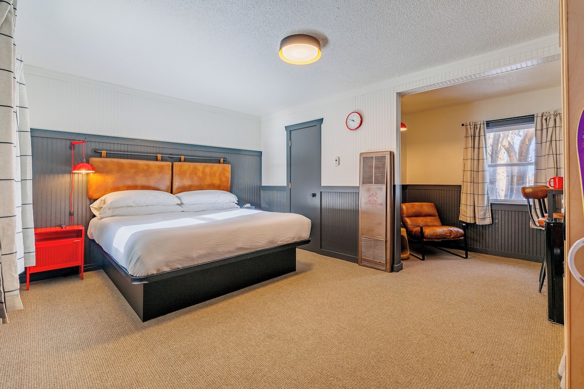 Ein Hotelzimmer mit einem Bett, einem roten Nachttisch, einem Stuhl, einem Heizgerät und einem Fenster mit Vorhängen.