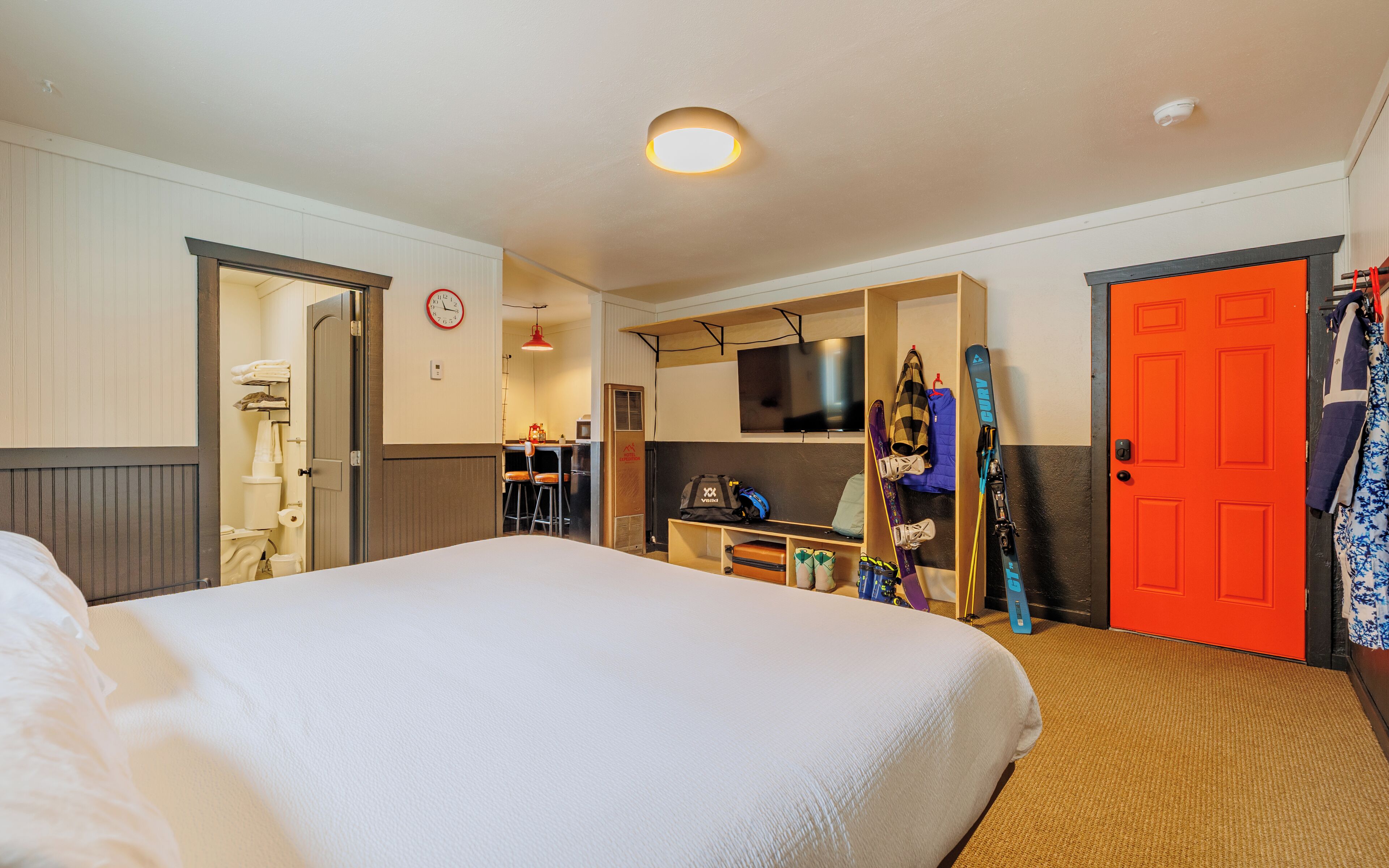 Une chambre d’hôtel avec un grand lit, un téléviseur fixé au mur, un porte-skis contenant des skis et du matériel, et une porte d’un orange vif.