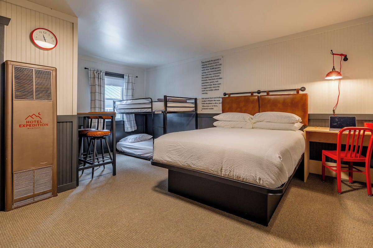 Ein Hotelzimmer mit Etagenbetten, einem Schreibtisch mit Stuhl, einem roten Sessel, einer Lampe, einer Uhr und einem Heizkörper.