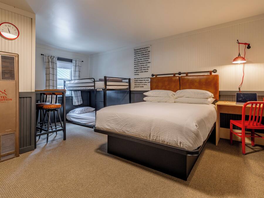 A hotel room with bunk beds, a desk with a chair, a red chair, a lamp, a clock, and a radiator.
