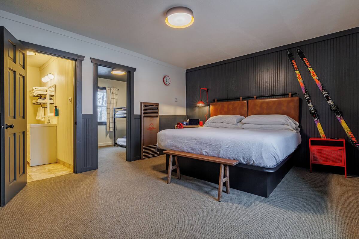 Ein modernes Hotelzimmer mit einem großen Bett, einem Schreibtisch und einer Bank. Ein roter Sessel und ein Skiständer mit Skiern sind ebenfalls vorhanden.