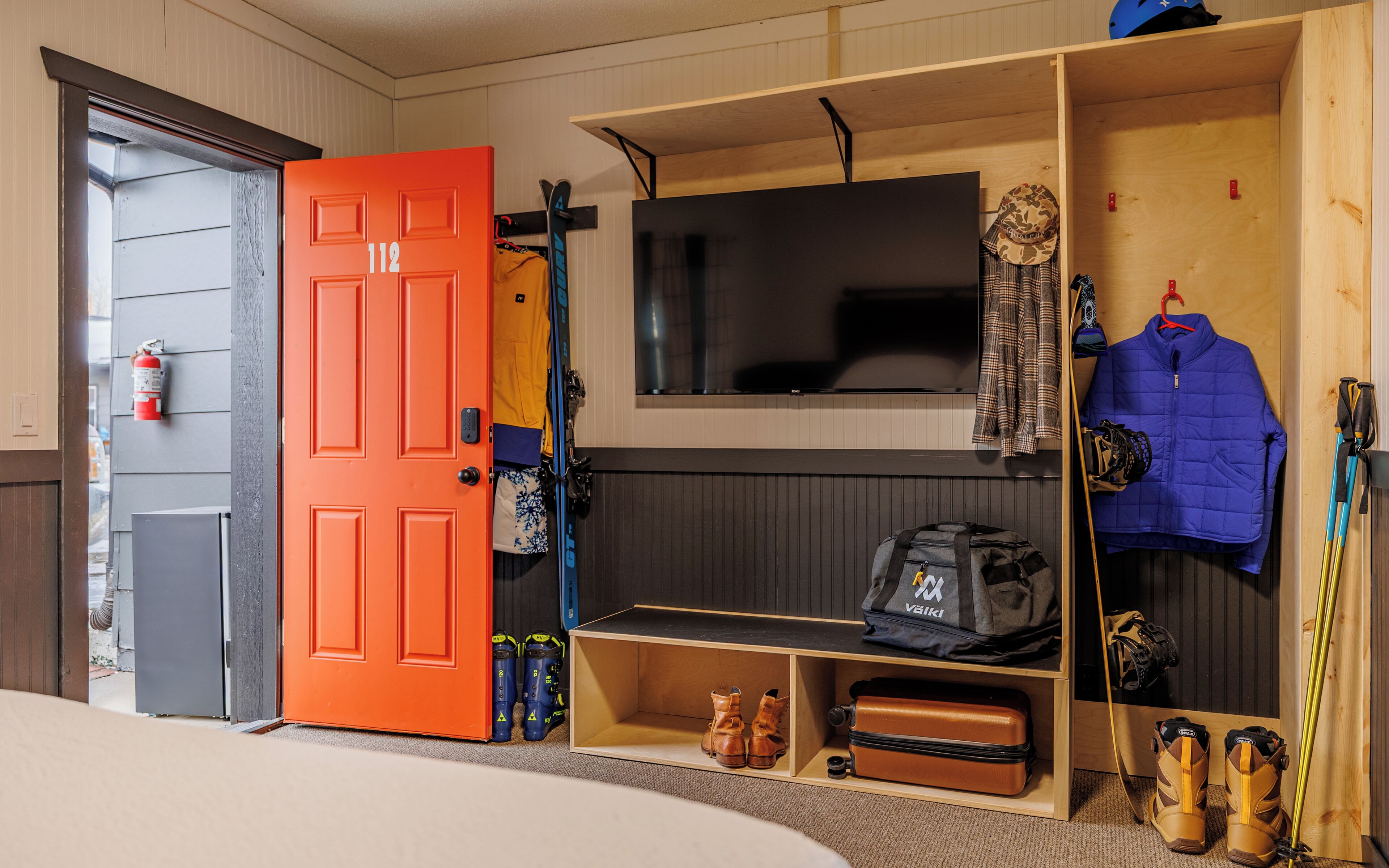 A room with a wooden bunk bed, a TV mounted on the wall, a fire extinguisher, and a suitcase.