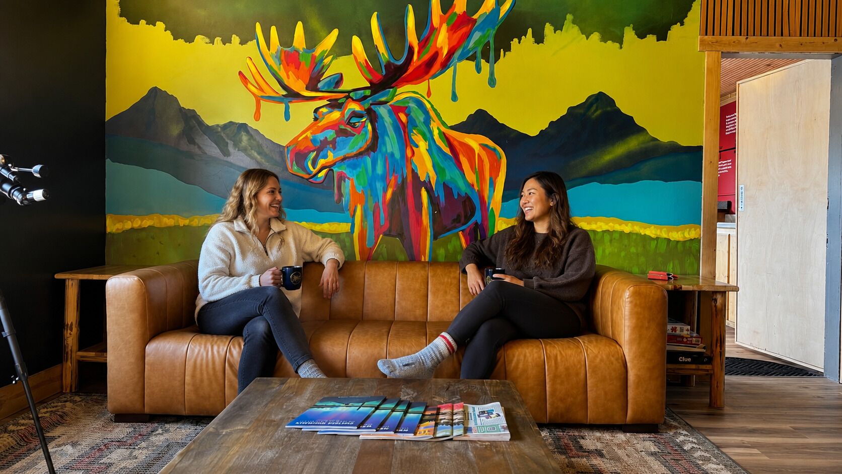 Two people sitting on a brown leather couch in a room with a colorful mural on the wall.