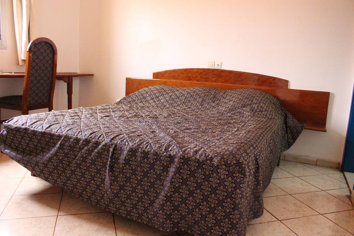 Un lit simple avec une couvre-lit à motifs, dans une chambre dotée d’une tête de lit en bois, d’une chaise et d’un bureau.