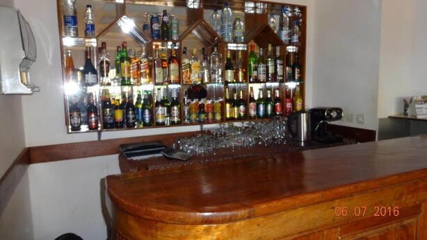 A bar area with a wooden counter, a mirrored wall displaying various bottles, and a coffee machine. - Hotel Altitel (Bafoussam)
