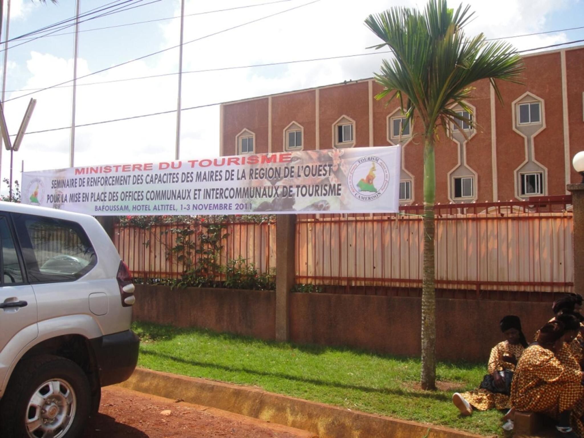 Un edificio con un cartel anunciando un seminario turístico, un automóvil blanco estacionado al frente y una palmera.