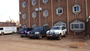 A building with a unique geometric facade, featuring multiple windows and air conditioning units. There are three vehicles parked in front, including a white van and two SUVs. - Hotel Altitel (Bafoussam)